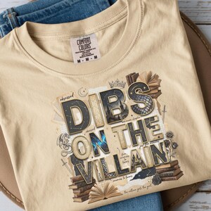 May include: Tan Comfort Colors t-shirt with the phrase "DIBS ON THE VILLAIN" in a stylized font, surrounded by illustrations of books, swords, and roses. The shirt is folded on a brown hat and blue jeans.