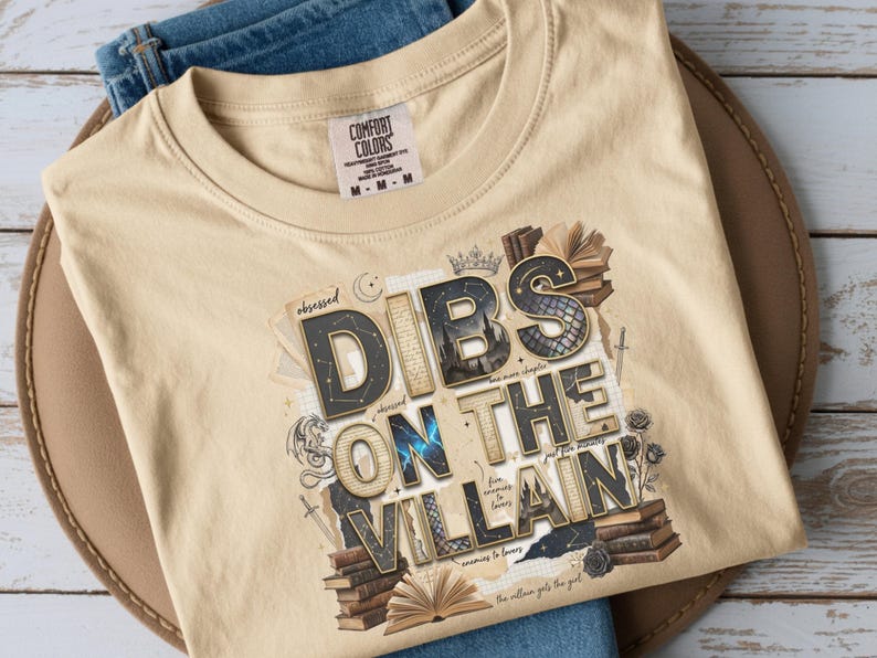 May include: Tan Comfort Colors t-shirt with the phrase "DIBS ON THE VILLAIN" in a stylized font, surrounded by illustrations of books, swords, and roses. The shirt is folded on a brown hat and blue jeans.
