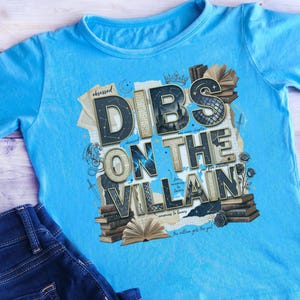 May include: A light blue t-shirt with the text "Dibs on the Villain" in a layered design. The graphic includes books, roses, and the word "Obsessed". A pair of blue jeans is visible in the image.