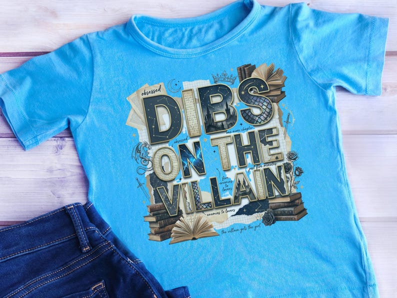 May include: A light blue t-shirt with the text "Dibs on the Villain" in a layered design. The graphic includes books, roses, and the word "Obsessed". A pair of blue jeans is visible in the image.