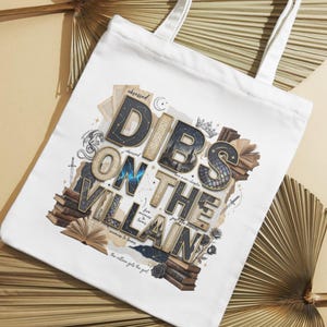 May include: White tote bag with the text "Dibs on the Villain" in a layered, vintage-style design. The design includes stacks of books, a crown, and other decorative elements. The bag has white handles.