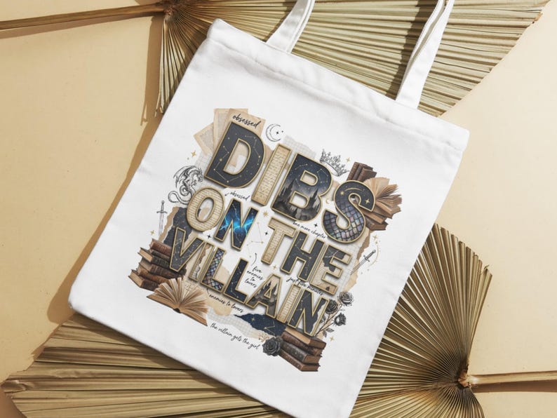 May include: White tote bag with the text "Dibs on the Villain" in a layered, vintage-style design. The design includes stacks of books, a crown, and other decorative elements. The bag has white handles.