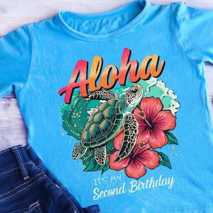 Puede incluir: Camiseta azul claro con un gráfico colorido. El diseño incluye la palabra "Aloha" en naranja y rojo, una tortuga marina, flores de hibisco y el texto "It's my Second Birthday". Ideal para un segundo cumpleaños.
