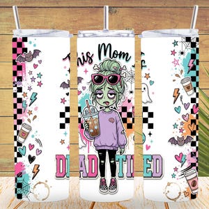 May include: A white cylindrical tumbler with a straw featuring a graphic of a zombie character with green hair and pink sunglasses holding an iced coffee. The text reads This Mom is DEAD TIRED, surrounded by bats, ghosts, hearts, and checkered patterns.
