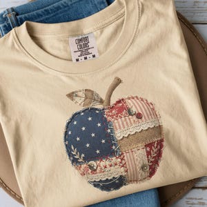 May include: A beige Comfort Colors t-shirt with a patchwork apple design. The apple incorporates star, stripe, and floral fabric patterns, with button and lace details. The shirt is folded on blue jeans. The shirt is folded on top of blue jeans.
