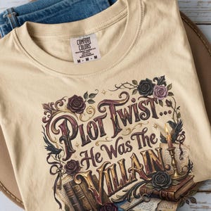 以下が含まれることがあります： 「Plot Twist... He Was The Villain」という文字がスタイリッシュなフォントでプリントされたベージュのTシャツ。デザインには、暗いバラ、真鍮の燭台に立てられた火のついたキャンドル、古い本、鍵のイラストが描かれています。