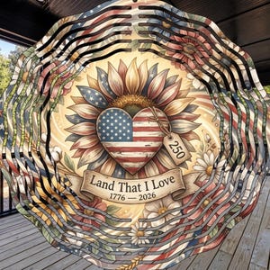 May include: A patriotic wind spinner featuring a sunflower with a heart-shaped American flag at its center. The design includes floral elements and the text "Land That I Love 1776-2026". The spinner has a layered, circular design.