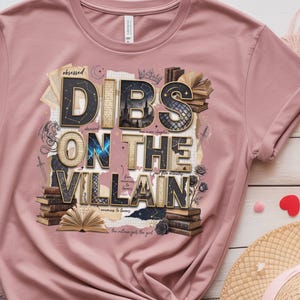 May include: A dusty rose t-shirt with the phrase "DIBS ON THE VILLAIN" in a layered, textured design. The design incorporates books, a crown, and a dragon illustration. The shirt is folded, with a straw hat and heart-shaped decorations in the background.