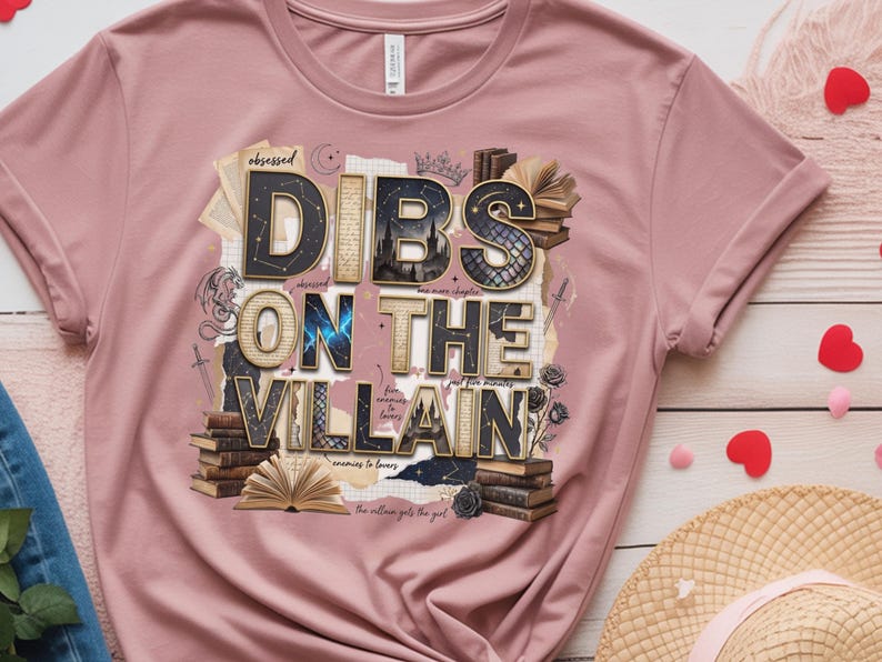 May include: A dusty rose t-shirt with the phrase "DIBS ON THE VILLAIN" in a layered, textured design. The design incorporates books, a crown, and a dragon illustration. The shirt is folded, with a straw hat and heart-shaped decorations in the background.