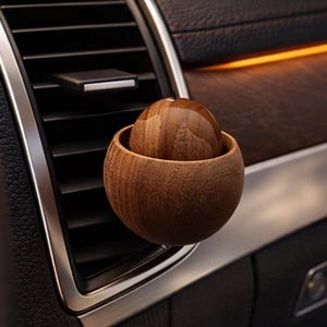 Puede incluir: Un ambientador de coche de madera con diseño esférico. El ambientador consta de una pequeña bola de madera dentro de un cuenco de madera más grande. El ambientador está colocado en una rejilla de ventilación del coche, con un interior oscuro y molduras de madera en el fondo.