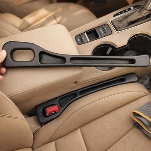 May include: Two black plastic car seat belt extenders. One extender is placed on a tan car seat, and the other is held by a hand. The extenders have a curved design with a slot for the seat belt buckle and a red release button.