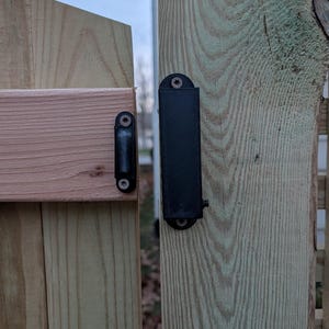 Op de afbeelding: Een houten poort met een zwarte metalen grendel en scharnier. De poort is gemaakt van lichtgekleurd hout met zichtbare nerf, en de grendel en het scharnier zijn met schroeven bevestigd. De poort staat gedeeltelijk open.