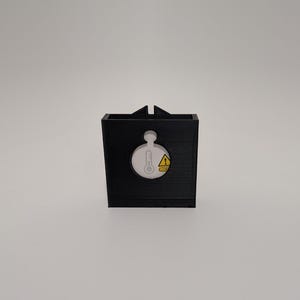 Può includere: Un contenitore nero stampato in 3D con un ritaglio circolare che mostra un termometro e un simbolo di avvertimento. Il contenitore ha una forma rettangolare con una piccola linguetta nella parte superiore. I simboli sono gialli e bianchi.