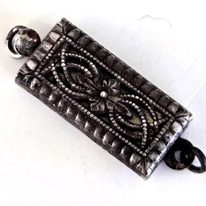 May include: A rectangular silver-toned metal jewelry clasp featuring an intricate embossed floral pattern and a beaded border. The clasp has a small circular loop attached to each of the two shorter ends, designed for connecting jewelry strands.