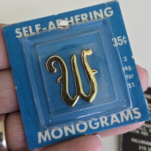 Puede incluir: Un monograma autoadhesivo dorado con la letra "W" en un paquete de plástico transparente sobre una tarjeta azul. La tarjeta dice "SELF-ADHERING MONOGRAMS" y "35¢". La letra tiene un estilo decorativo y cursivo.