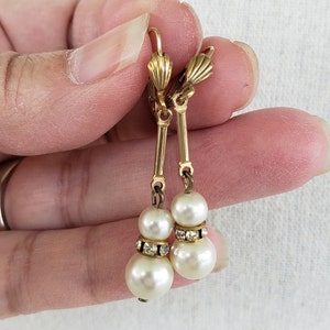 Puede incluir: Pendientes de gota de tono dorado con dos cuentas de perla blanca y detalles de pedrería transparente.