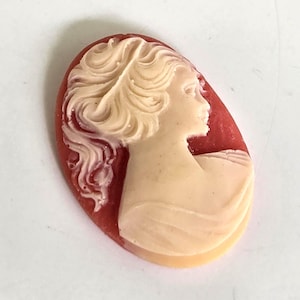 May include: Oval cameo pendant featuring a profile of a woman with flowing hair. The woman's profile is a light cream color, set against a reddish-brown background. The cameo is a classic, vintage-inspired piece.