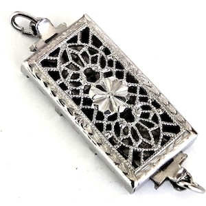 May include: A rectangular silver-toned metal pendant featuring an intricate openwork filigree design with a central floral motif. The frame is decorated with fine engraved patterns and includes a small jump ring at both the top and bottom ends.
