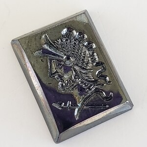 May include: A rectangular, iridescent, silver-coloured metal button with a raised relief design. The design features a stylized figure with a feathered headdress, spear, and ornate details. The button has a slightly bevelled edge.