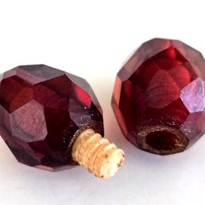 May include: Two faceted, deep red, translucent beads with a glossy finish. One bead features a protruding, light-colored, threaded screw-like attachment at its base, while the other bead displays a circular opening at the center of its base.
