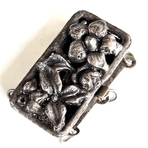 May include: A silver-toned rectangular jewelry clasp with a floral design. The clasp features a raised relief of flowers and leaves within a rectangular frame. The clasp has three loops for attachment and a small metal bar for closure.