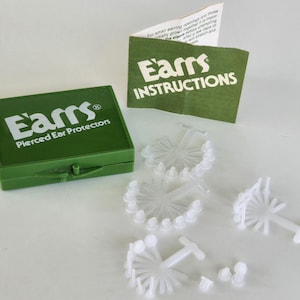 May include: A green box with the text "Earns Pierced Ear Protectors" on the lid. The box is open and contains a variety of white plastic ear protectors. There is also a separate instruction sheet with the text "Earns Instructions" on it.