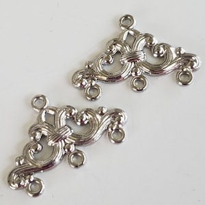 May include: Two silver-colored filigree connectors with ornate, swirling designs. Each connector features multiple loops for attaching to other components. The connectors have a symmetrical, decorative appearance, suitable for jewelry making or craft projects.