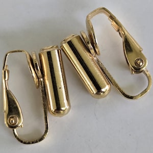 Peut inclure: Gros plan sur quatre apprêts de boucles d'oreilles dorées. Chaque apprêt est composé d'une tige cylindrique avec une fermeture à levier articulée. Les apprêts sont en matériau métallique brillant, sur fond blanc.