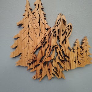 May include: Wooden wall art depicting a howling wolf silhouette amidst a forest of pine trees. The artwork is crafted from light brown wood with intricate cut-out details, creating a layered effect. The wolf is positioned in the center, with trees on either side.