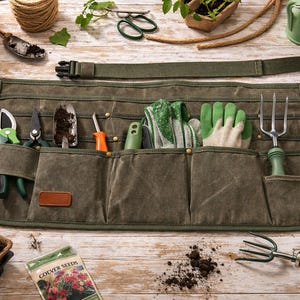 Pode incluir: Avental de jardinagem de lona verde com vários bolsos, contendo diversas ferramentas como tesouras, espátulas e um garfo de mão. O avental está sobre uma superfície de madeira, com suprimentos de jardinagem adicionais e plantas ao fundo.