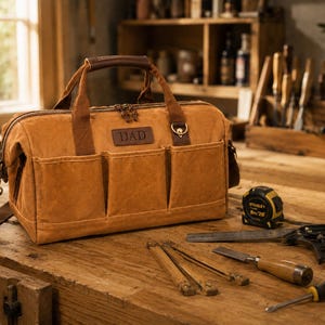 May include: A tan canvas tool bag with brown leather handles and trim, featuring three exterior pockets and a rectangular leather patch embossed with the word DAD. The bag includes a detachable shoulder strap, brass hardware, and a top zipper closure.