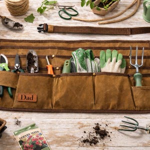 May include: A brown canvas gardening apron with pockets, holding tools such as shears, trowels, and gloves. The apron has a leather patch that says "Dad". Other gardening tools and supplies are arranged around the apron.