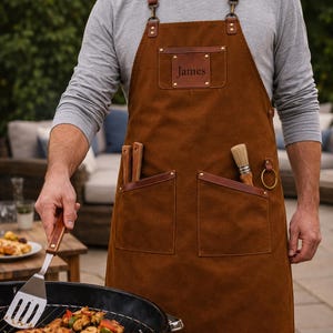 May include: A brown canvas apron featuring leather straps with metal buckles, a leather chest patch embossed with the name James, two large front pockets, and a side loop. The apron is worn over a grey long-sleeve shirt while grilling food.