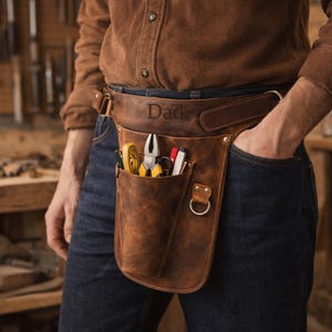 May include: A brown leather tool belt with the word "Dad" embossed on the front. The belt contains tools such as pliers, a tape measure, and a pen. The belt is worn by a person wearing a brown shirt and blue jeans.