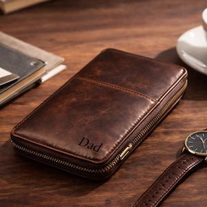 Könnte beinhalten: Eine braune Ledergeldbörse mit dem eingravierten Wort "Dad", eine goldfarbene Uhr mit braunem Lederarmband, eine Tasse Kaffee und ein Notizbuch mit Stift auf einer Holzoberfläche.