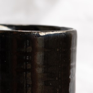 May include: Close-up of a dark brown ceramic mug with a glossy finish. The rim has a slightly uneven texture with a lighter colour. The mug's surface reflects light, highlighting its shape and texture.