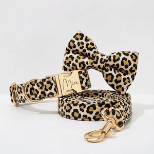 Puede incluir: Un conjunto de accesorios para mascotas con estampado de leopardo, que incluye un collar, una correa y una pajarita. El collar tiene una hebilla dorada grabada con el nombre "Mia". La correa tiene un mosquetón dorado. El conjunto es marrón y negro.