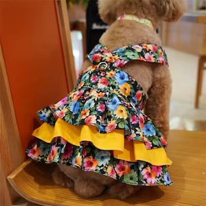 Pode incluir: Vestido para cães com estampa floral em fundo preto, com flores rosa, azuis e amarelas. O vestido tem uma saia em camadas com uma camada amarela brilhante e um detalhe de laço no decote.