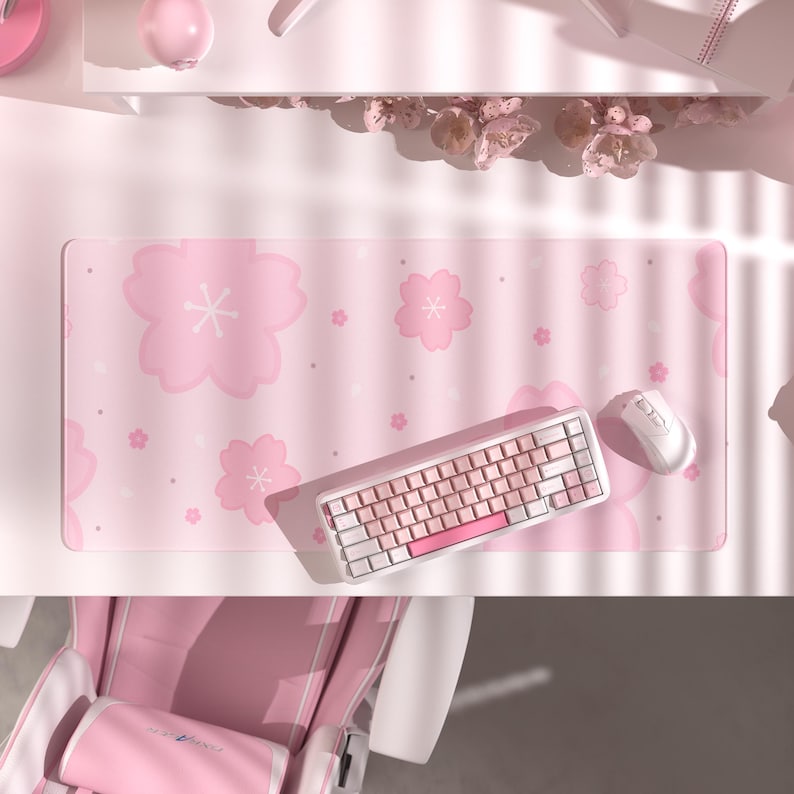 K&ouml;nnte beinhalten: Ein rosa Schreibtisch-Setup mit einem Mauspad mit Blumenmuster, einer wei&szlig;en Tastatur mit rosa Akzenten und einer wei&szlig;en Maus. Das Setup befindet sich auf einem wei&szlig;en Schreibtisch, darunter ist ein rosa Stuhl zu sehen.