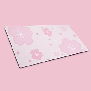 K&ouml;nnte beinhalten: Rechteckige, rosa Schreibtischunterlage mit einem sich wiederholenden Muster aus stilisierten rosa Kirschbl&uuml;ten mit wei&szlig;en Zentren. Kleine wei&szlig;e und graue Punkte sind &uuml;ber dem hellrosa Hintergrund verstreut. Die Matte hat einen schwarzen Rand.