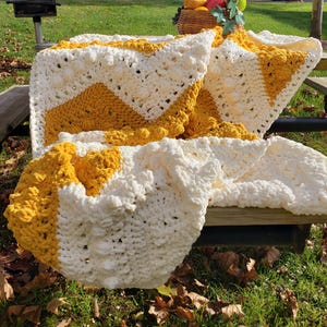May include: A handmade, chunky knit blanket in white and mustard yellow. The blanket features a chevron design and is draped over a wooden picnic table. The blanket is made of a soft, textured yarn, perfect for snuggling up on a chilly day.