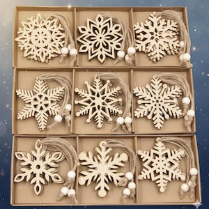 Peut inclure: Neuf décorations en forme de flocons de neige en bois dans une boîte en carton. Chaque flocon de neige a un design unique et est attaché à une boucle de ficelle avec des perles blanches. Ornements de couleur bois naturel.