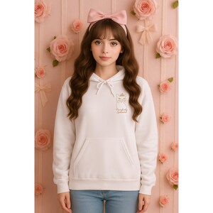 May include: White hoodie with a front pocket and drawstring. The hoodie features a gold outline of a cat wearing a crown, with the text "Purrfectly Splendid" below. The person is wearing blue jeans and a pink bow headband.