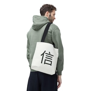 Puede incluir: Un bolso tote blanco con una correa negra para el hombro y un gran carácter japonés negro. El bolso se lleva al hombro. La persona de la imagen lleva una sudadera con capucha verde.