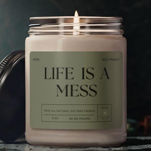 Könnte beinhalten: Eine brennende Sojawachskerze in einem durchsichtigen Glas mit silbernem Deckel und schwarzem Schraubverschluss. Auf dem Etikett steht "LIFE IS A MESS" in schwarzer Schrift auf grünem Hintergrund. Die Kerze hat 170g und brennt 50-60 Stunden lang.