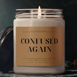 Puede incluir: Una vela de cera de soja encendida en un tarro de cristal transparente con tapa negra. La etiqueta dice "CONFUSED AGAIN" en negrita. La vela es de 255 g y tiene un tiempo de combustión de 50-60 horas.