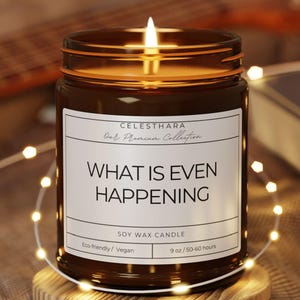 May include: A lit soy wax candle in a brown glass jar with the text "WHAT IS EVEN HAPPENING" on a white label. The candle sits on a small wooden coaster, surrounded by string lights. The candle is 9 oz and burns for 50-60 hours.
