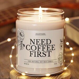 May include: A lit soy wax candle in a glass jar with the text "NEED COFFEE FIRST" on a label. The candle sits on a round wooden coaster. The candle is 9 oz and has a burn time of 50-60 hours.