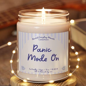 Könnte beinhalten: Eine brennende Kerze in einem Glas mit der Aufschrift "Panic Mode On". Die Kerze steht auf einem kleinen Holzuntersetzer, umgeben von Lichterketten. Im Hintergrund sind eine Gitarre und ein Buch zu sehen.