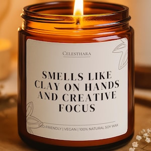 May include: A lit soy wax candle in a brown glass jar with a white label. The label reads "Smells Like Clay on Hands and Creative Focus." The candle sits on a wooden tray, with a blurred light and plant in the background.